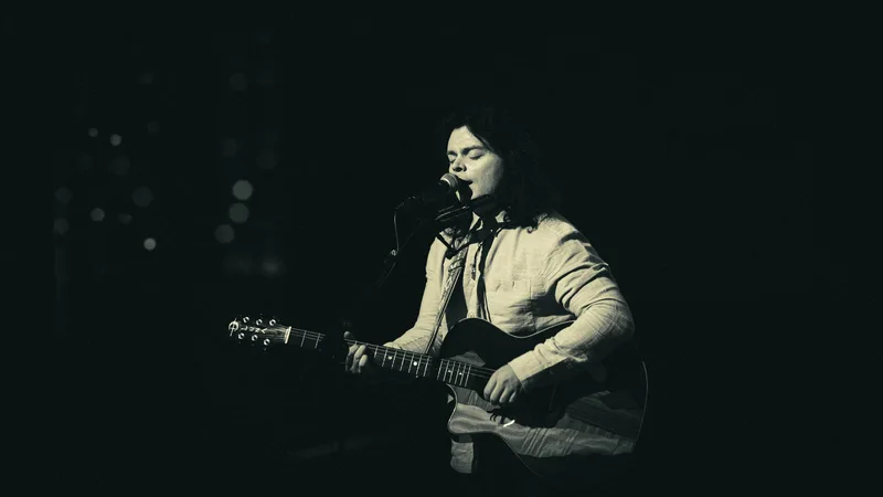 Black and white frame of a singer leaning into the microphone, guitar in hand.