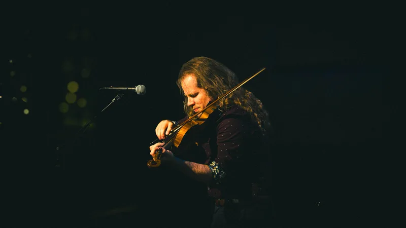 A violinist in a dark shirt draws the bow, lit warm against a black stage.
