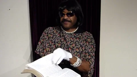 A person dressed in drag reading a book while pointing at the text.
