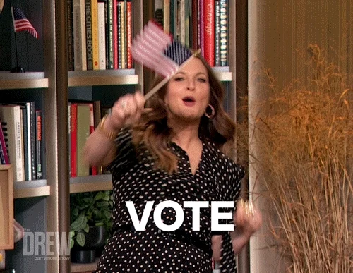 Drew Barrymore standing in a library, holding a US flag and boxing in the air with both arms saying, 