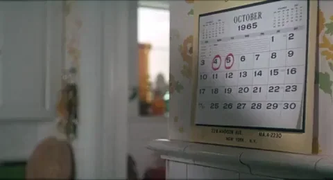 A woman looking at a calendar of October on the wall with a red pen in her mouth.