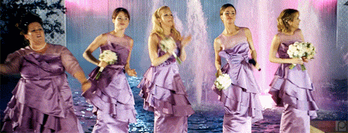 5 women in purple dresses from movie Bridesmaids dancing by a fountain.
