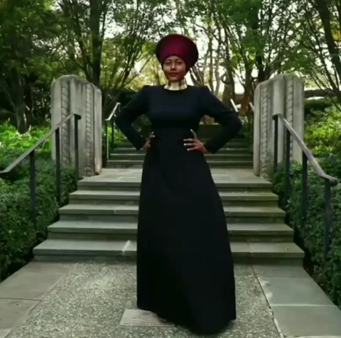 A Black woman in traditional clothing, posing in front of stone stairs in a public garden. The text reads, 'African Queen'.