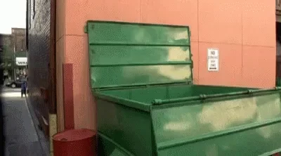 A man in blue jeans is being thrown into a big green industrial garbage container.