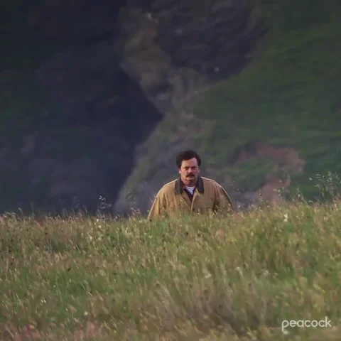 Nick Offerman walking up a grassy field. The camera zooms out to reveal a scenic landscape.