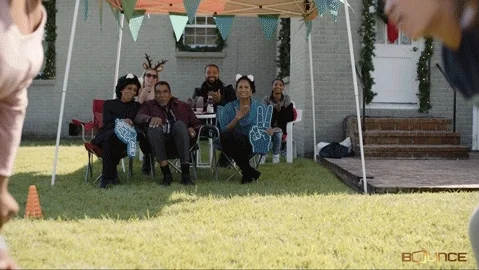 A family gathering with some members playing football and others cheering them on from under an awning in the backyard.