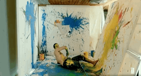 A shirtless artist in a studio. He whips a paint-covered shirt into a canvas, leaving a large paint streak on the canvas.