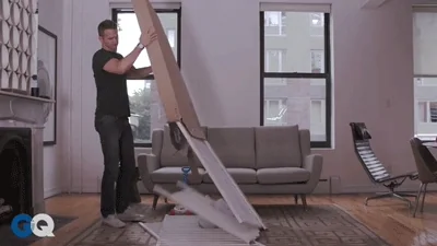 A man is moving a box with furniture pieces. They all fall out onto the floor. 