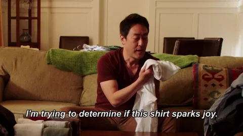 A man sitting on a couch and holding a white t-shirt, saying: