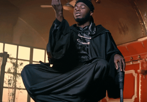 A man sitting on a chair wearing a black robe. His eyes are closed. He brings his hand down and gathers his fingers.
