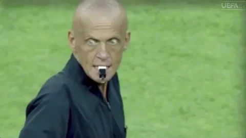 A soccer referee with wild eyes blows a whistle. 