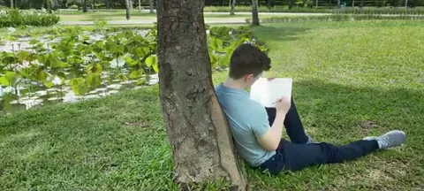 A man leaning against a tree stump while sitting in the grass under the tree shade, and writing in a journal.