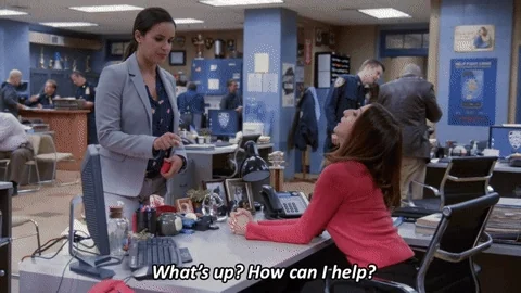 Gina from Brooklyn 99, wearing a red turtleneck, leans over a desk and asks, 