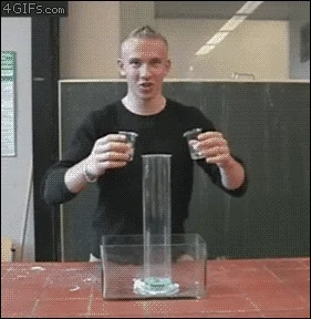 A man pours liquids from two small beakers into a larger tall container. Quickly the reaction foams, shooting up. 