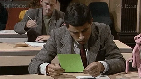 Mr. Bean sitting in a classroom reviewing a paper during an exam, covering his eyes after reading it.