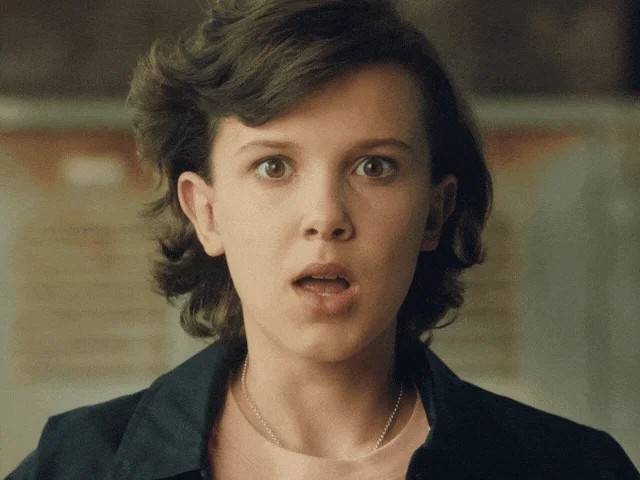 Eleven from Stranger Things says “Boom!” while making an exploding gesture with her hands from the side of her face.