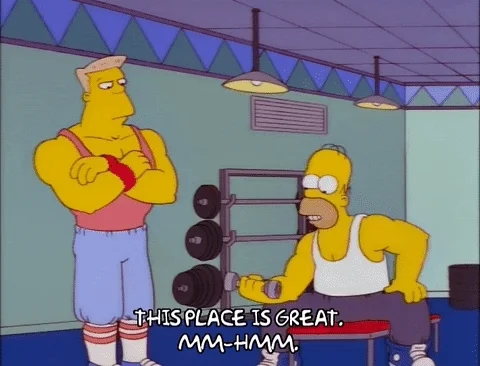 Homer Simpson lifting a small hand weight while a buff trainer looks on. He's saying, 'This place is great, mm-hmm.'