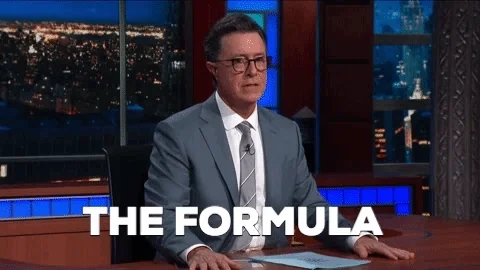 Stephen Colbert, sitting at a desk, wearing a grey suit jacket and white shirt with grey striped tie saying, 