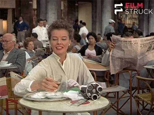 A woman by herself having a drink at a cafe in an old movie.