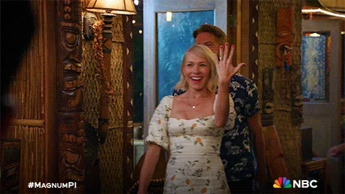 A blonde woman in a white floral dress walks into a room holding up her hand with an engagement ring on it. 