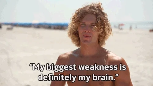 A shirtless man with long curly hair is standing on a beach and saying 