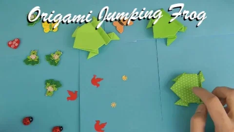 A GIF of a person's hand testing origami frogs jumping on a blue surface, with the overlay text reading Origami Jumping Frog.