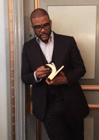 Tyler Perry wearing stylish suit, leaning against a door, and flipping through a book.
