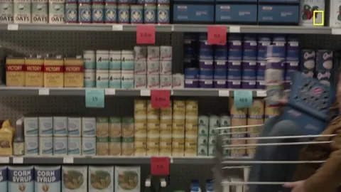 One person is pushing another in a cart in front of shelves filled with food.