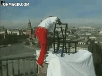An acrobat balancing on a stacked chairs at the top of a building.