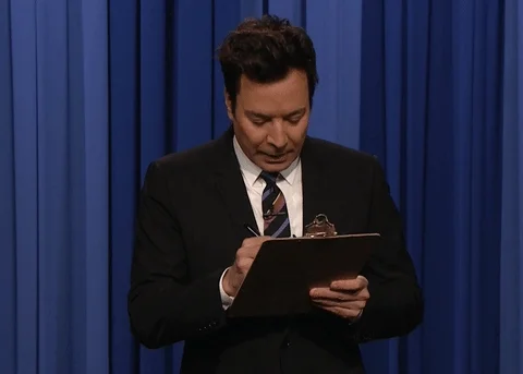 Jimmy Fallon on stage taking notes on a clipboard.
