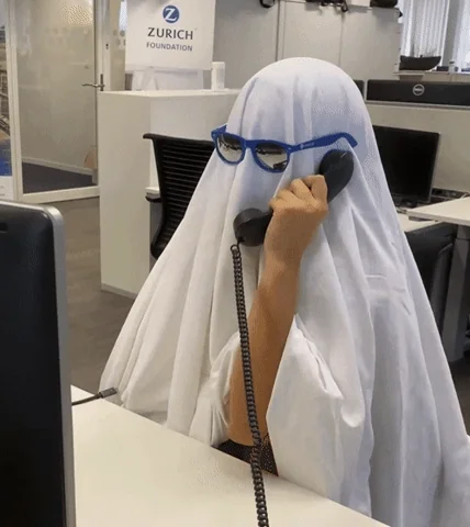 A person disguised as a ghost in a bed sheet wearing blue glasses, sitting at an office desk and talking on the phone.