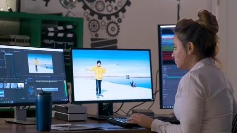 A woman sits at multiple computer screens doing video editing