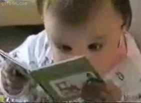 Baby holding small book up closely to her face, moving head back and forth as if she's reading.
