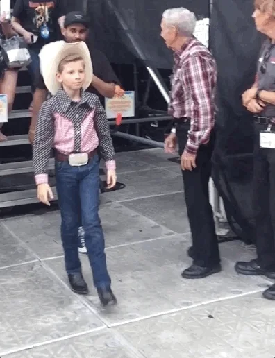 A child dressed as cowboy does a little dance walking past others and ends with a twirl and fingers pointing out. 