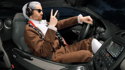 A man wearing sunglasses and headphones sits in the driver's seat of a car, holding up a peace sign with his hand.