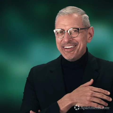 Man with gray hair, dark clothes, and dark glasses smiling and pointing finger at you saying, 