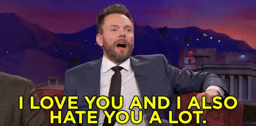 Joel McHale is sitting on a couch on a talk show stage with a mountain background shaking his head. 
