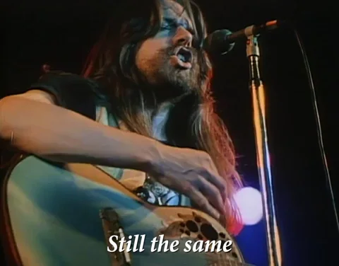 A man with long hair playing a guitar and singing on stage. Text says: 'Still the same'.