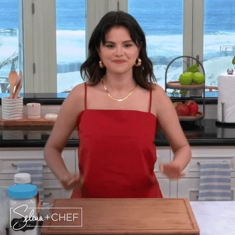 Selena Gomez in front of a cutting board, with a red apron on, giving two thumbs up while saying, 