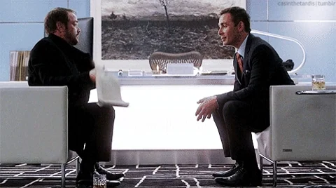 Two men sitting together, as one of them unfurls a long scroll filled with text.