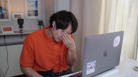 A man sits in front of a laptop, pinching the bridge of his nose as if he has a headache.