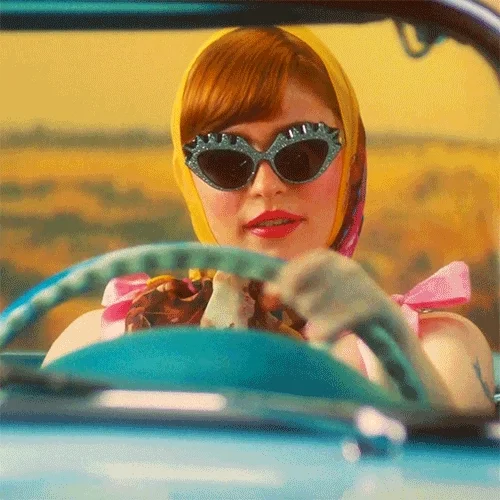 A woman wearing blue sunglasses and a shawl drives a vintage car across a desert landscape.