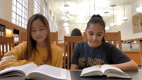 Two girls are sitting at a table in a library and giving each other a high five. There are open books on the table.  
