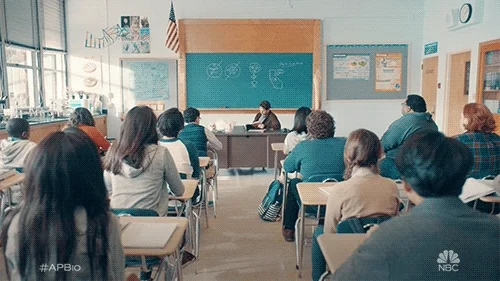 A few students in a high school classroom anxiously raise their hands while a teacher looks down at their desk. 