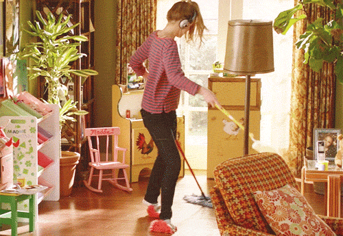 Woman wearing headphones and dancing to music while mopping, dusting and cleaning the floor with cloths under her feet.