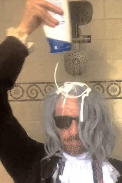A man wearing an 18th century costume and standing in a shower pours shampoo on his wig.