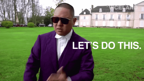A man in a suit and sunglasses on the grounds of a mansion. He says, 'Let's do this.'