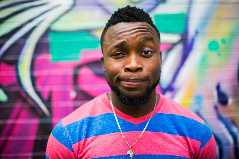 An African American man standing in front of a graffiti wall, wearing a pink and blue striped shirt, making a silly face.