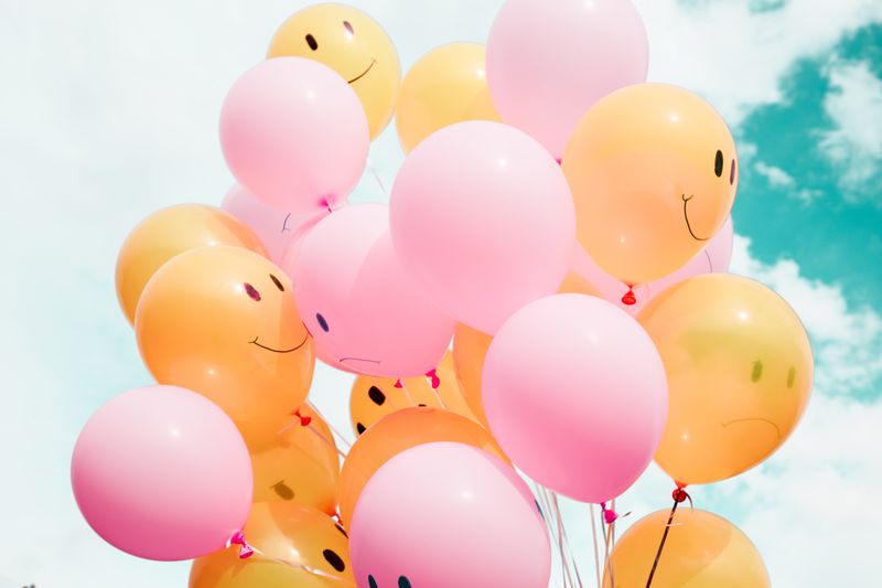 A bunch of yellow and pink balloons with smiling faces drawn on them floating into a sunny blue sky with white puffy clouds.