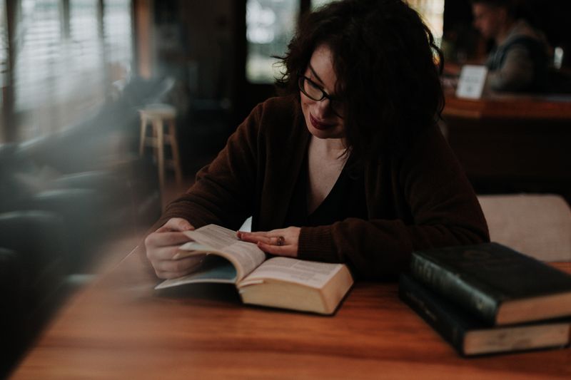 A young woman with dark hair and glasses is reading a book in a cafe.